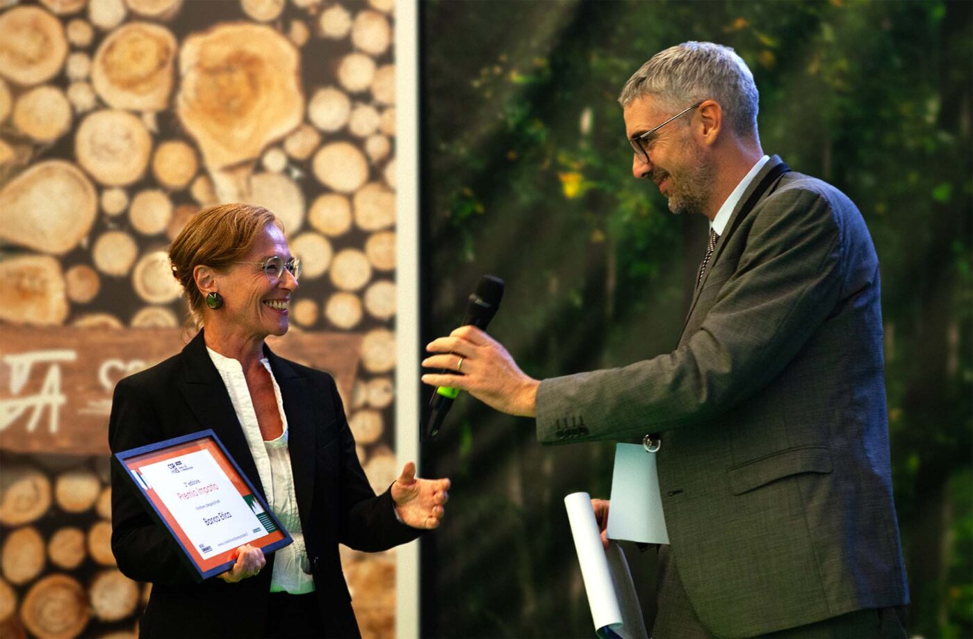 Two professionals interact, with one receiving a microphone and the other holding an award.