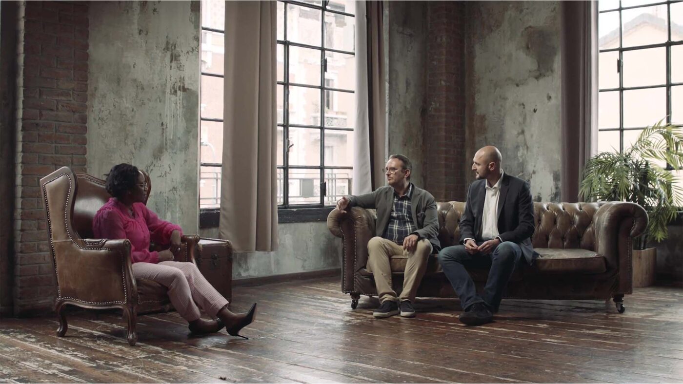 Three people are seated in a spacious room with large windows, engaged in conversation. One person is in an armchair, two on a sofa.
