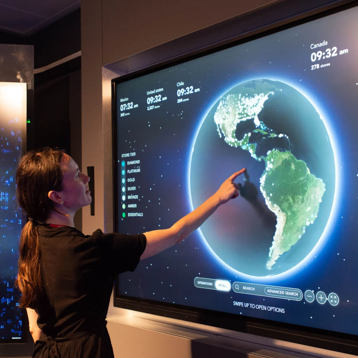 A person points at a large interactive screen displaying a digital globe with store locations and time zones