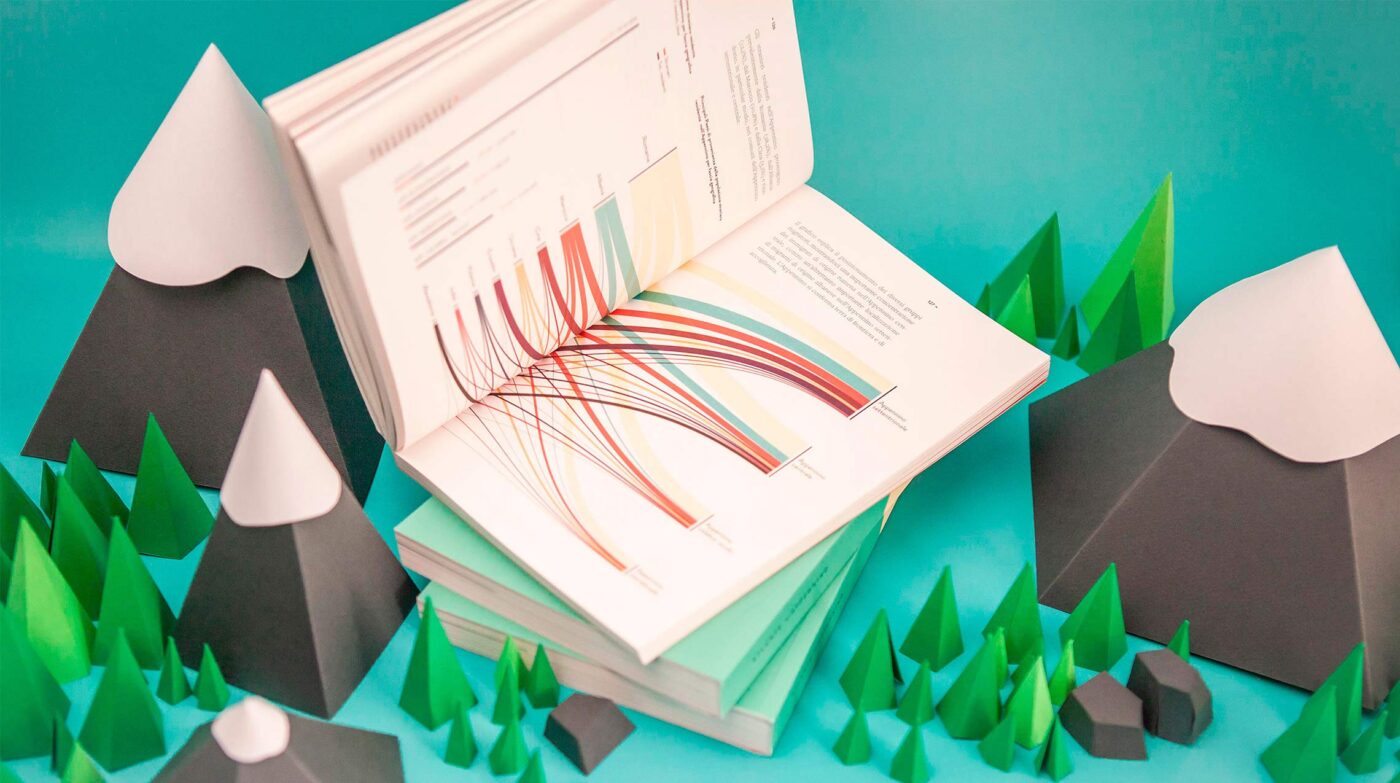 An open book displays a colorful data visualization with flowing lines, surrounded by a miniature paper landscape of mountains and trees.